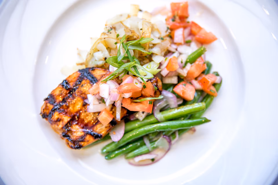 A plated meal featuring a grilled piece of salmon topped with diced tomatoes and onions, served with sautéed green beans and caramelized onions on a white plate.