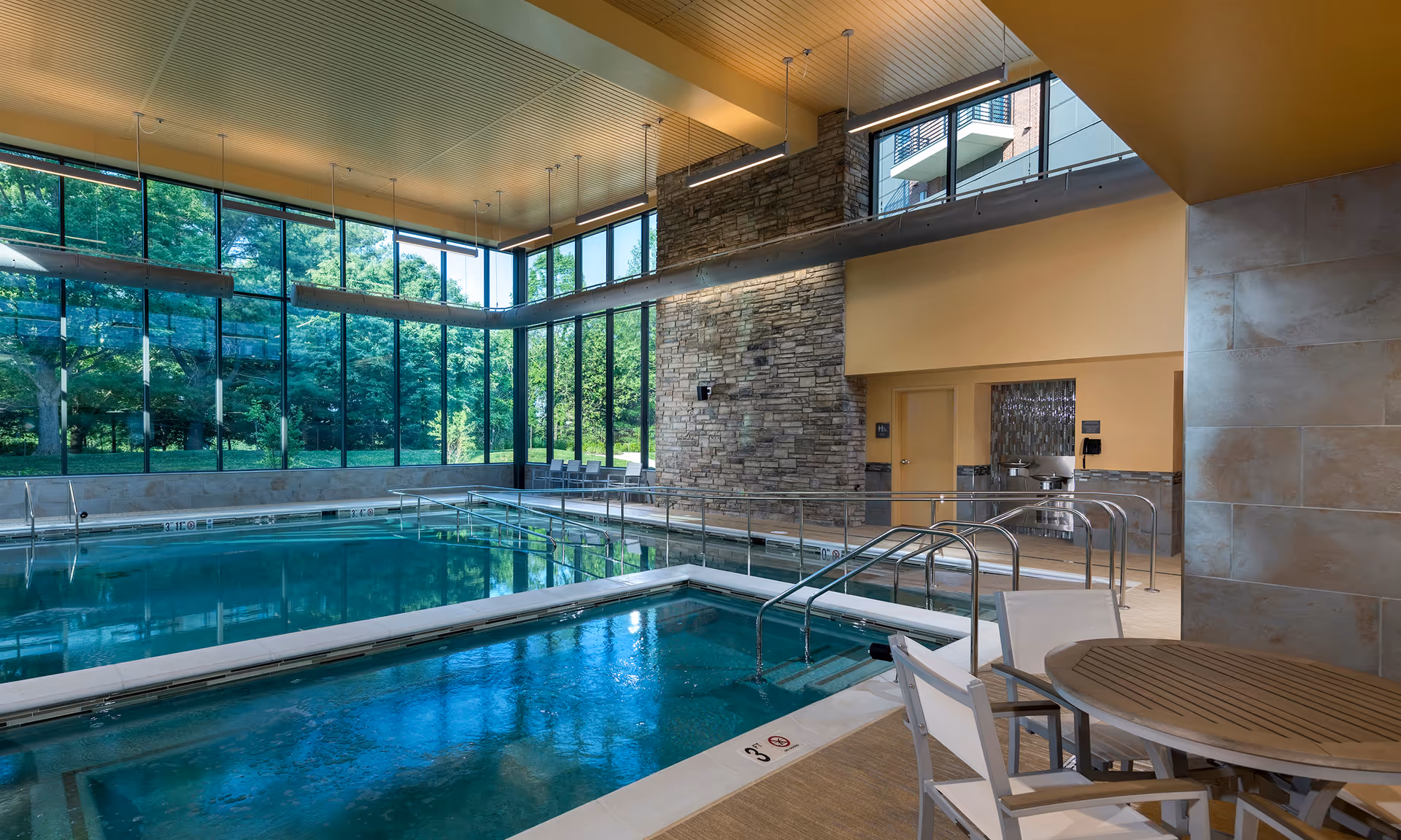 Indoor swimming pool area with floor-to-ceiling windows, a stone feature wall, pool handrails and seating with tables and chairs.