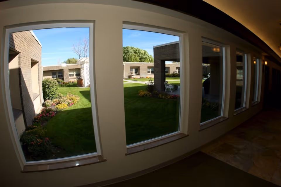 A curved indoor corridor with large windows overlooking a landscaped courtyard with grass, flowerbeds, and single-story buildings.