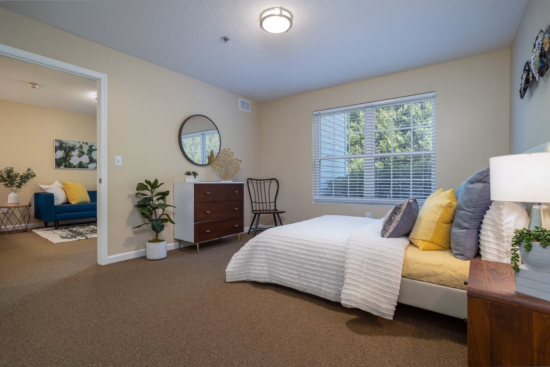 A bright bedroom with a large window letting in natural light. The bed is made with white and yellow bedding and several pillows. Next to the bed is a wooden nightstand with a lamp and a small plant. Across from the bed is a dresser with a round mirror above it, a decorative coral piece, and a potted plant on the floor. A black chair is placed near the window. An open doorway leads to a living area with a blue sofa, yellow pillows, a side table with a plant, and a wall art piece featuring white flowers.