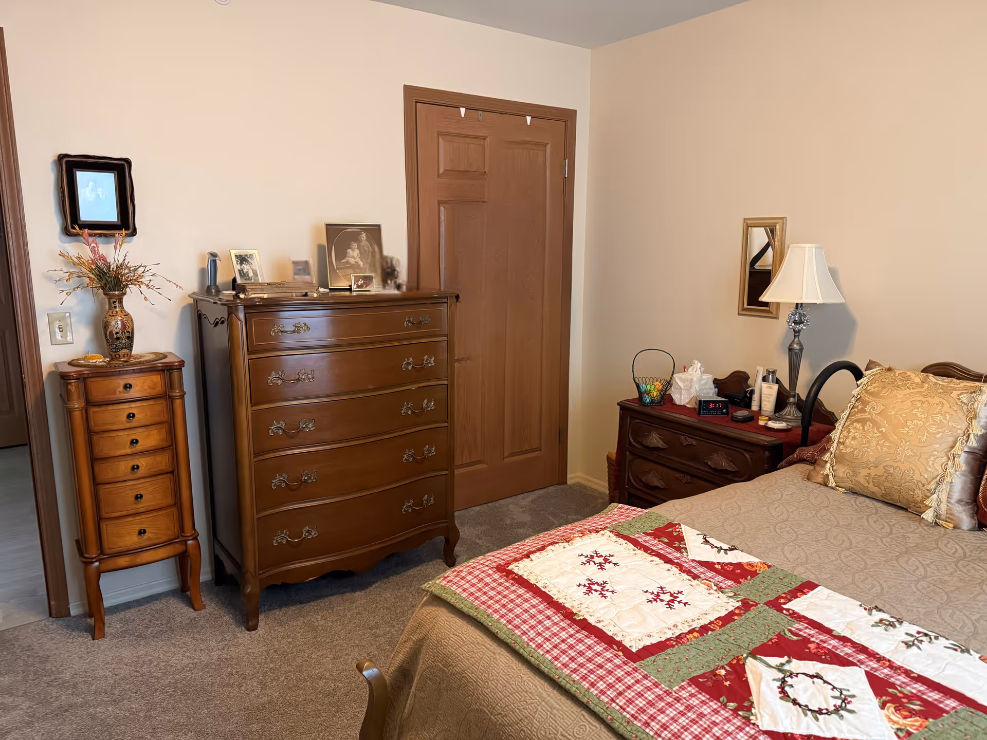 A cozy bedroom with a quilted bed, wooden dressers and nightstand with a lamp, and a closed wooden door.