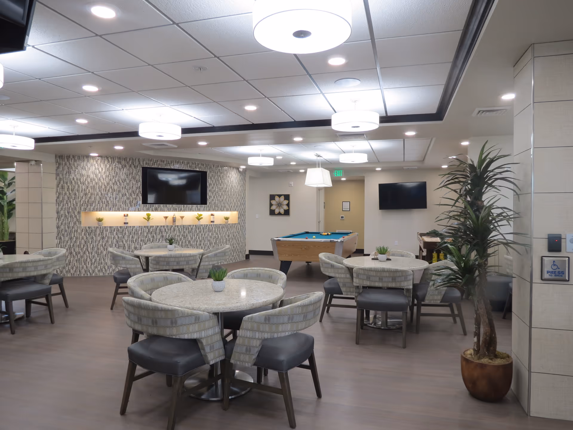 Spacious senior living common room with round dining tables and chairs, a pool table, wall-mounted TVs, and potted plants.