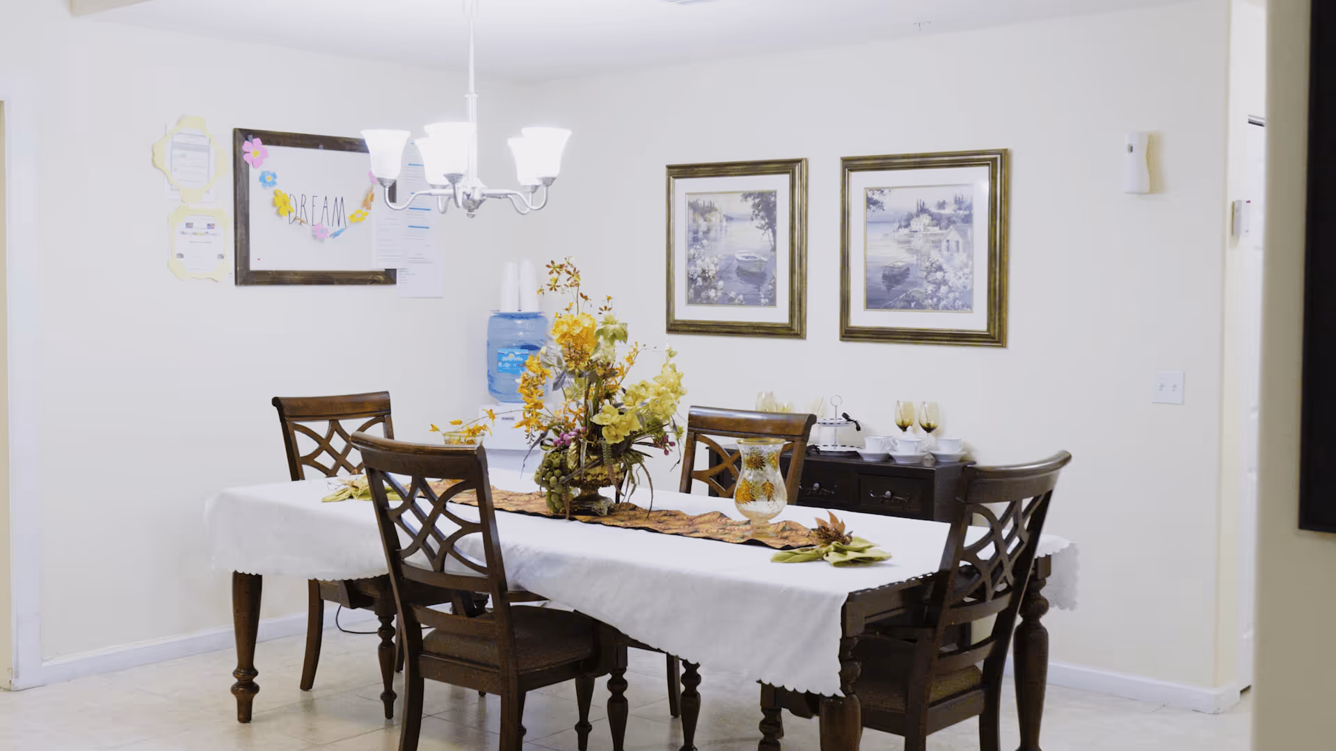 Dining room with a rectangular table covered by a white tablecloth, wooden chairs, a floral centerpiece, chandelier, framed artwork on the wall and a water cooler.