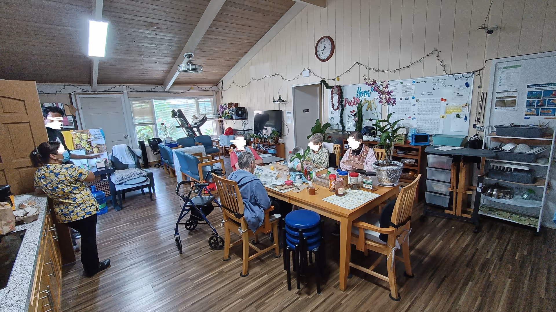 A group of elderly people sitting around a wooden table in a communal room with various items on the table including plants, jars, and papers. A caregiver stands nearby wearing a mask and a colorful shirt. The room has wooden floors, a high ceiling with exposed beams, a clock on the wall, a whiteboard with notes, and several chairs and exercise equipment near the windows.