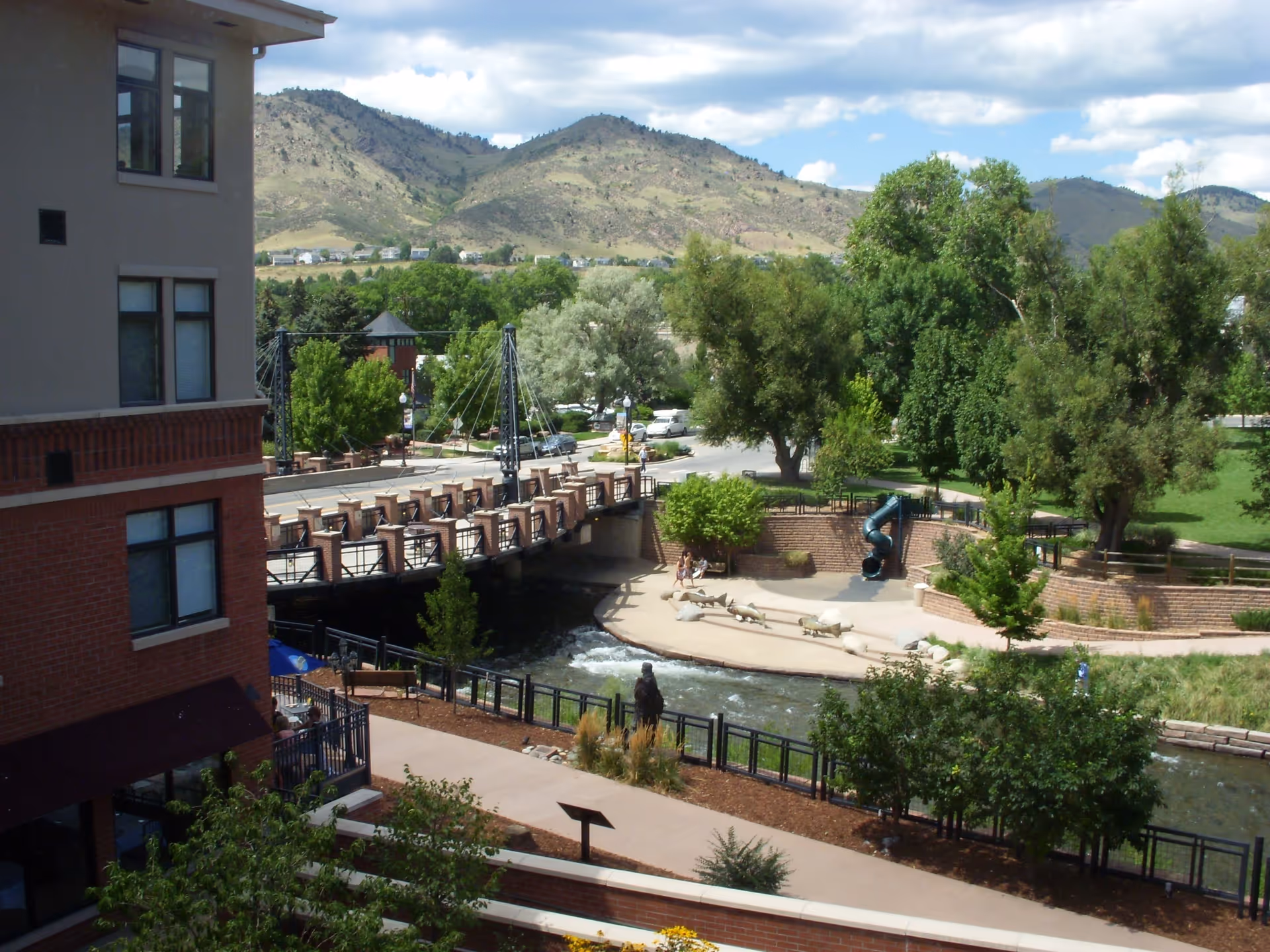 View of a riverside park with a pedestrian bridge, walkways, trees, a small slide, and an adjacent brick building with hills in the background.
