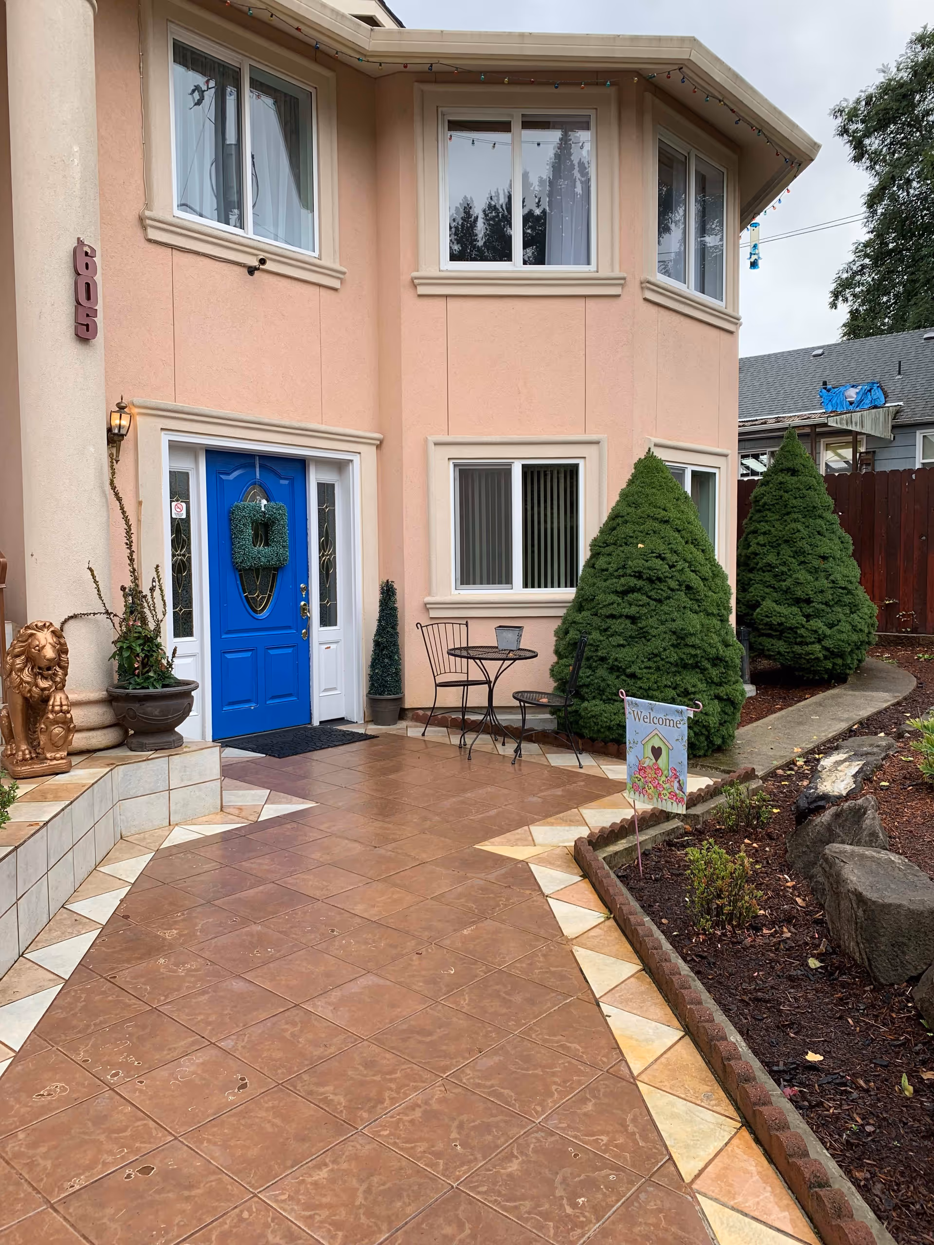 Front entrance of a two-story building with a bright blue door decorated with a green wreath. The building has peach-colored walls and several windows with white frames. There is a tiled walkway leading to the door, flanked by two large green bushes and a small garden area with rocks and plants. A small round metal table with two chairs is placed near the window. A decorative lion statue and potted plants are near the entrance. A small sign in the garden reads 'Welcome'.