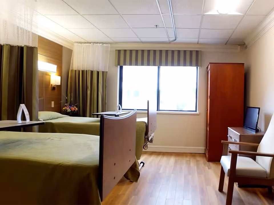 A bright and clean room with two hospital-style beds covered in green bedspreads, separated by a curtain. The room has a large window with striped valance, a wooden wardrobe, a small TV on a dresser, and a white armchair with wooden arms. The floor is wooden, and the walls are light-colored.