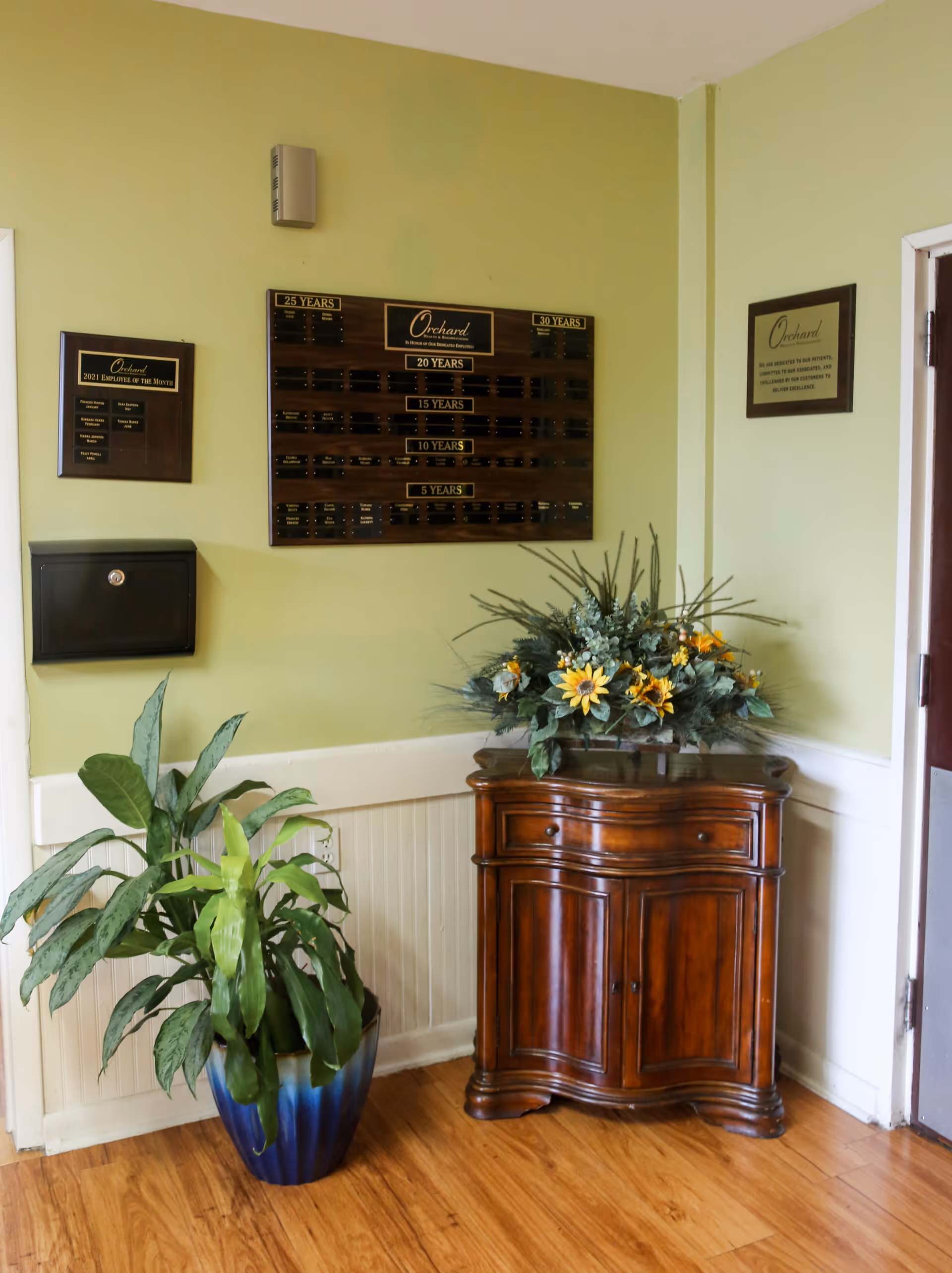 Corner of a room with light green walls featuring a wooden plaque honoring years of service, a smaller plaque for employee of the month, and a framed sign. Below the plaques is a dark wooden cabinet with a floral arrangement on top and a blue and green potted plant on the floor beside it. The floor is wooden, and a door is partially visible on the right.