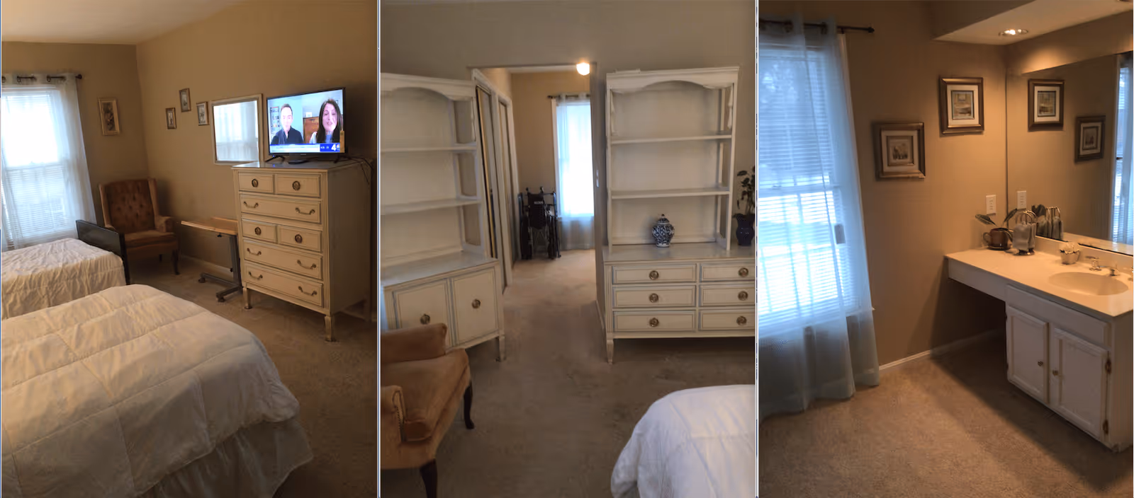 A senior living facility bedroom with two twin beds covered in white comforters, a beige armchair, a white dresser with a TV on top, and a small overbed table. The room has beige walls and carpet, with windows covered by sheer curtains. Adjacent to the bedroom is a vanity area with a large mirror, sink, and countertop, decorated with framed pictures on the wall and small decorative items on the counter.