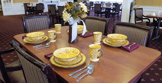 A dining table set for four with yellow patterned plates, bowls, mugs, and silverware. A floral centerpiece with white and yellow flowers is in the middle of the table. The room has multiple similar tables and chairs with striped upholstery and a carpeted floor.