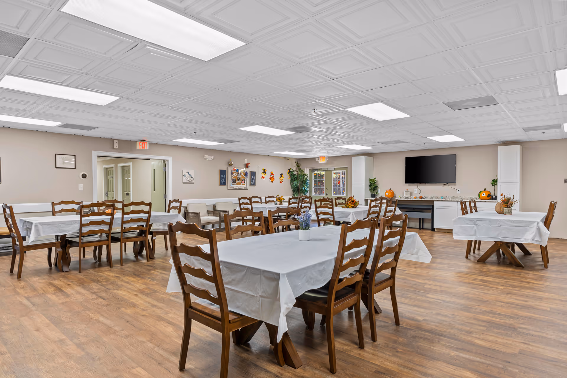 A spacious dining room with multiple wooden tables and chairs covered with white tablecloths. The room has a wooden floor, beige walls, and a white ceiling with fluorescent lights. There are decorations on the walls and tables, including small plants and seasonal ornaments. A large flat-screen TV is mounted on the far wall above a countertop with cabinets.