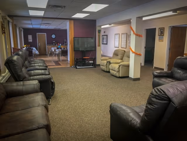 A common area in a senior living facility with several leather recliners arranged along the walls. In the background, there is a flat-screen TV on a stand and a dining area with tables and chairs. The room has carpeted flooring and fluorescent ceiling lights.