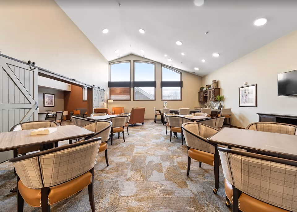 A spacious senior living dining room with multiple tables and cushioned chairs arranged neatly. The room features high vaulted ceilings with recessed lighting, large windows with blinds allowing natural light, and a cozy seating area with armchairs in the back. There is a cabinet with decorative items and a wall-mounted TV on the right side.