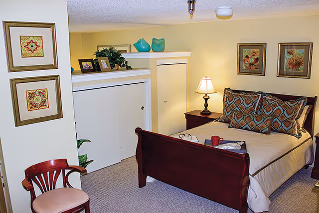 A cozy bedroom with a wooden bed frame, beige bedding, and patterned pillows. There is a nightstand with a lamp and a red cup on the bed. The room has framed artwork on the walls, a wooden chair with a cushioned seat, and decorative items on a shelf.