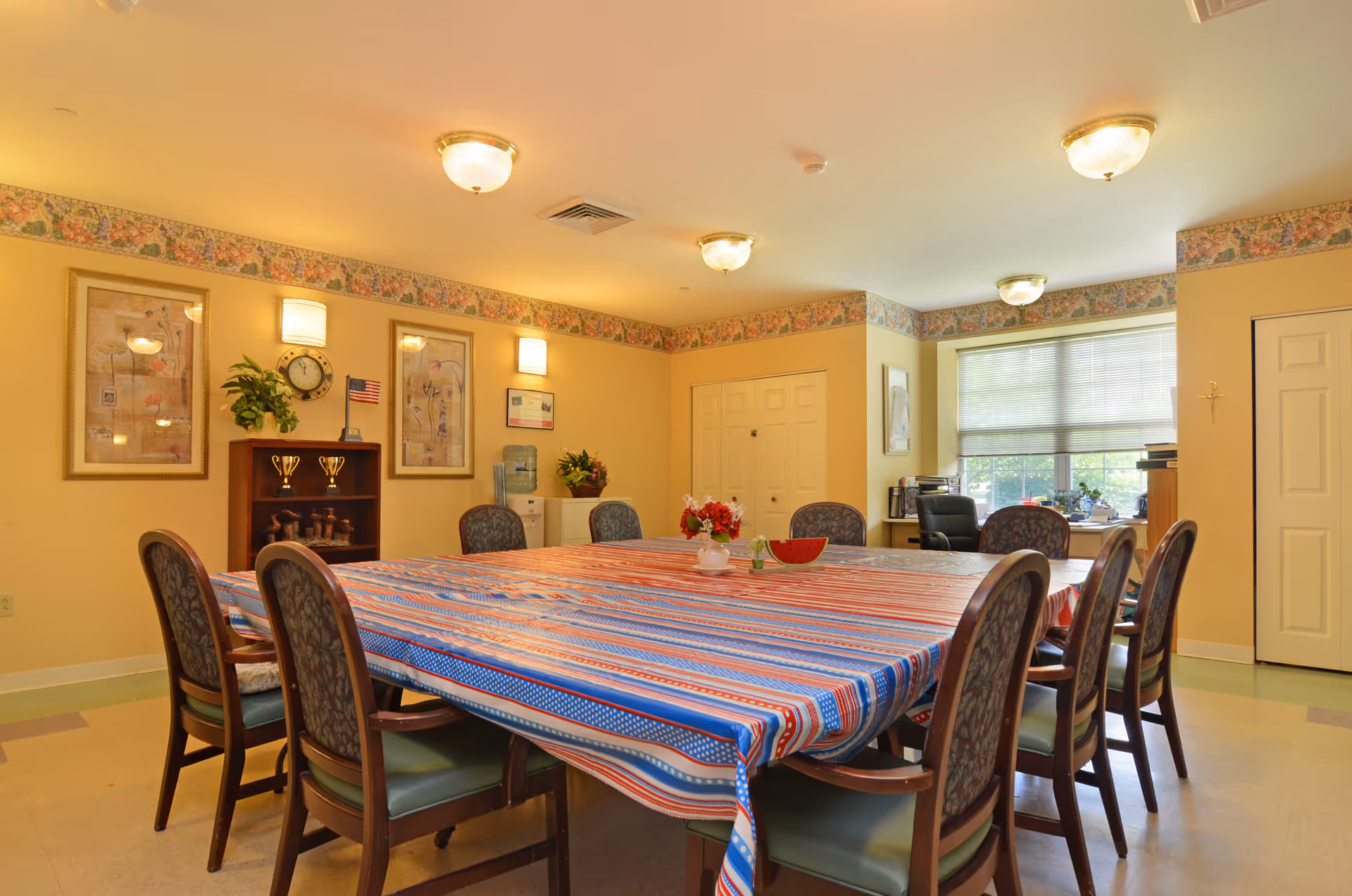 A well-lit dining room with a large rectangular table covered with a red, white, and blue striped tablecloth. The table is surrounded by cushioned chairs with floral upholstery. The room has yellow walls with floral border wallpaper near the ceiling, framed artwork, a clock, and a small American flag on a shelf. There is a water cooler and a window with blinds letting in natural light.