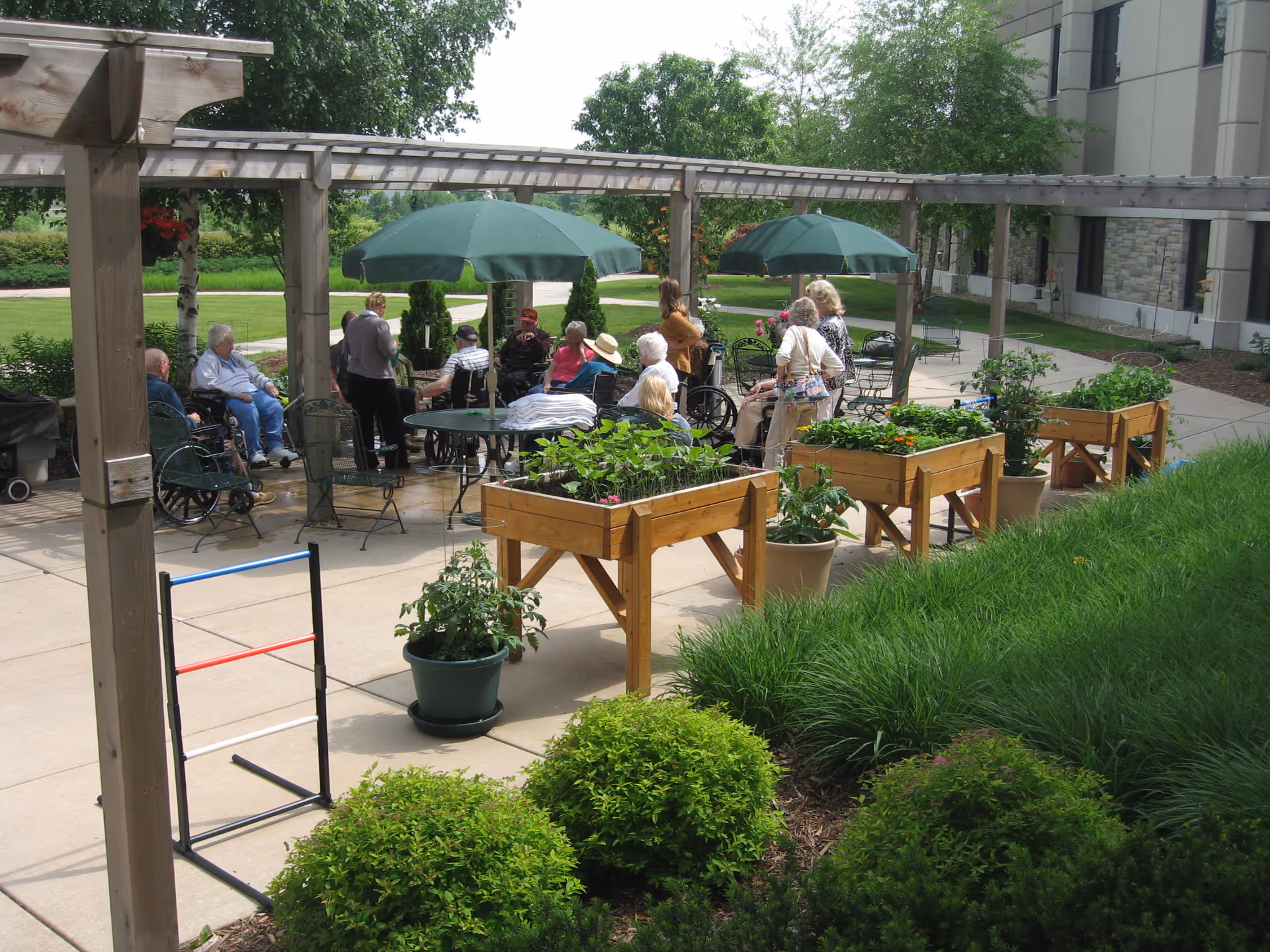 A group of elderly people sitting and standing under a wooden pergola with green umbrellas on a patio outside a building. There are raised garden beds with plants and flowers, potted plants, and green bushes surrounding the patio area.