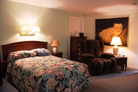 A cozy bedroom with a floral-patterned bedspread on a wooden bed frame, a nightstand with a lamp, a cushioned recliner chair, a wooden dresser, and a large framed picture of an orange cat on the wall.