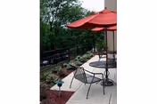 Outdoor patio with metal tables and chairs under red umbrellas next to landscaped planting beds and a fence.