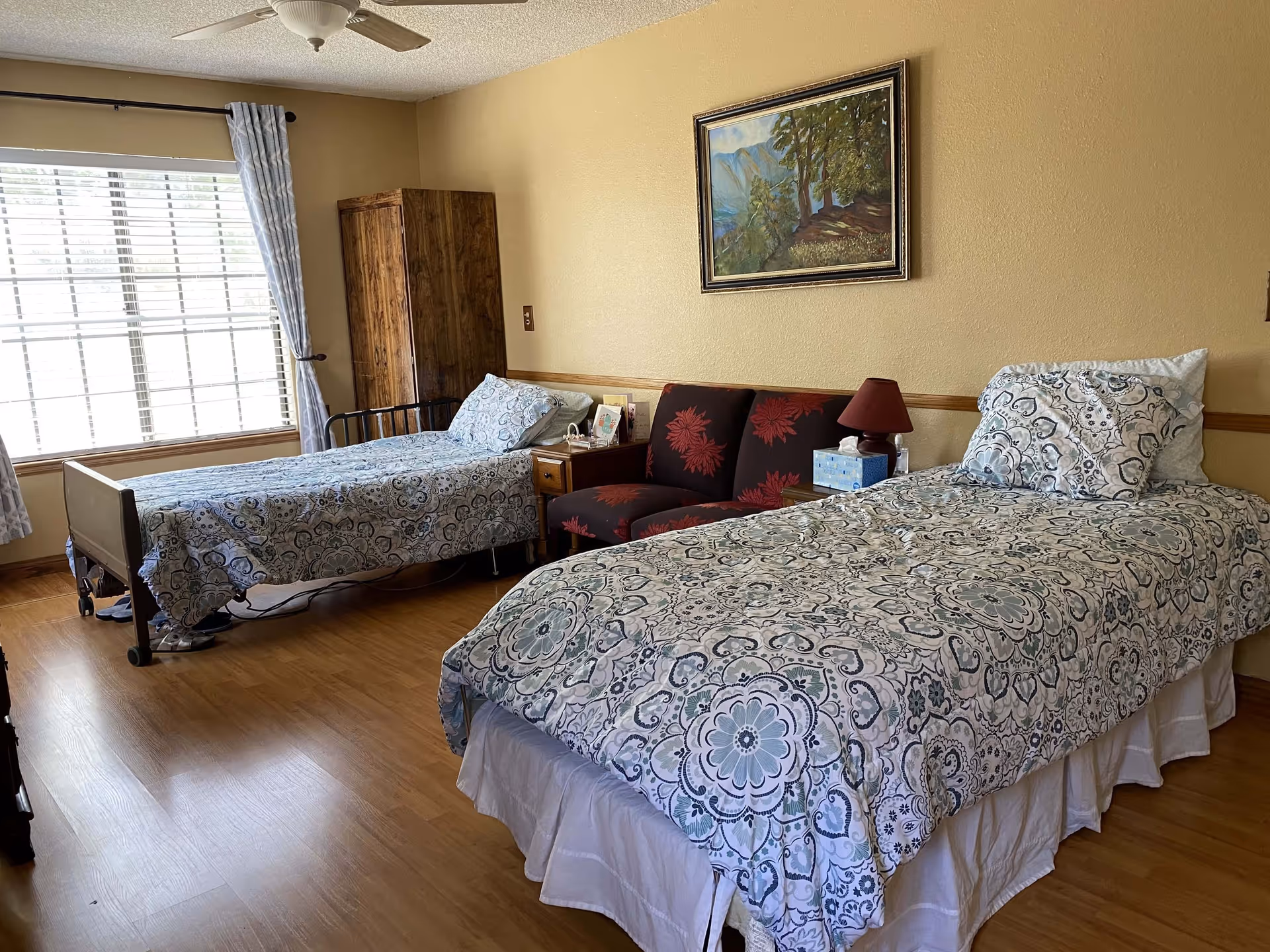 A bedroom with two single beds covered in matching patterned bedspreads. Between the beds is a small wooden nightstand with a red lamp, tissue box, and hand sanitizer. A small red loveseat with floral patterns is placed against the wall behind the nightstand. There is a large window with light blue curtains on the left side and a wooden wardrobe in the corner. A framed landscape painting hangs on the beige wall above the loveseat.