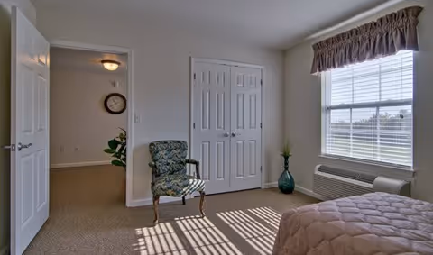 Sunny furnished bedroom with a bed, patterned chair, double-door closet, and a window with blinds.