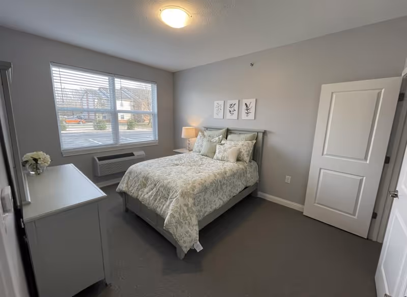 A neatly arranged bedroom with a bed covered in floral-patterned bedding, a bedside table with a lamp, a dresser with a small flower arrangement, a large window with blinds showing a view of houses outside, and a closed white door.