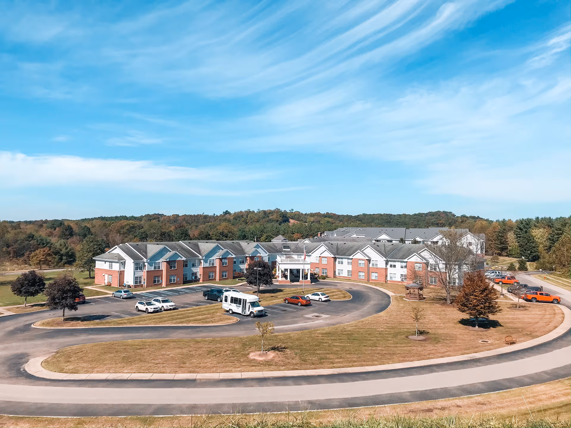 Aerial view of The Glenwood Community senior living facility showing a large two-story building with a circular driveway and parking lot. Several cars and a shuttle bus are parked outside. The building is surrounded by grassy areas, trees, and a backdrop of forested hills under a partly cloudy blue sky.