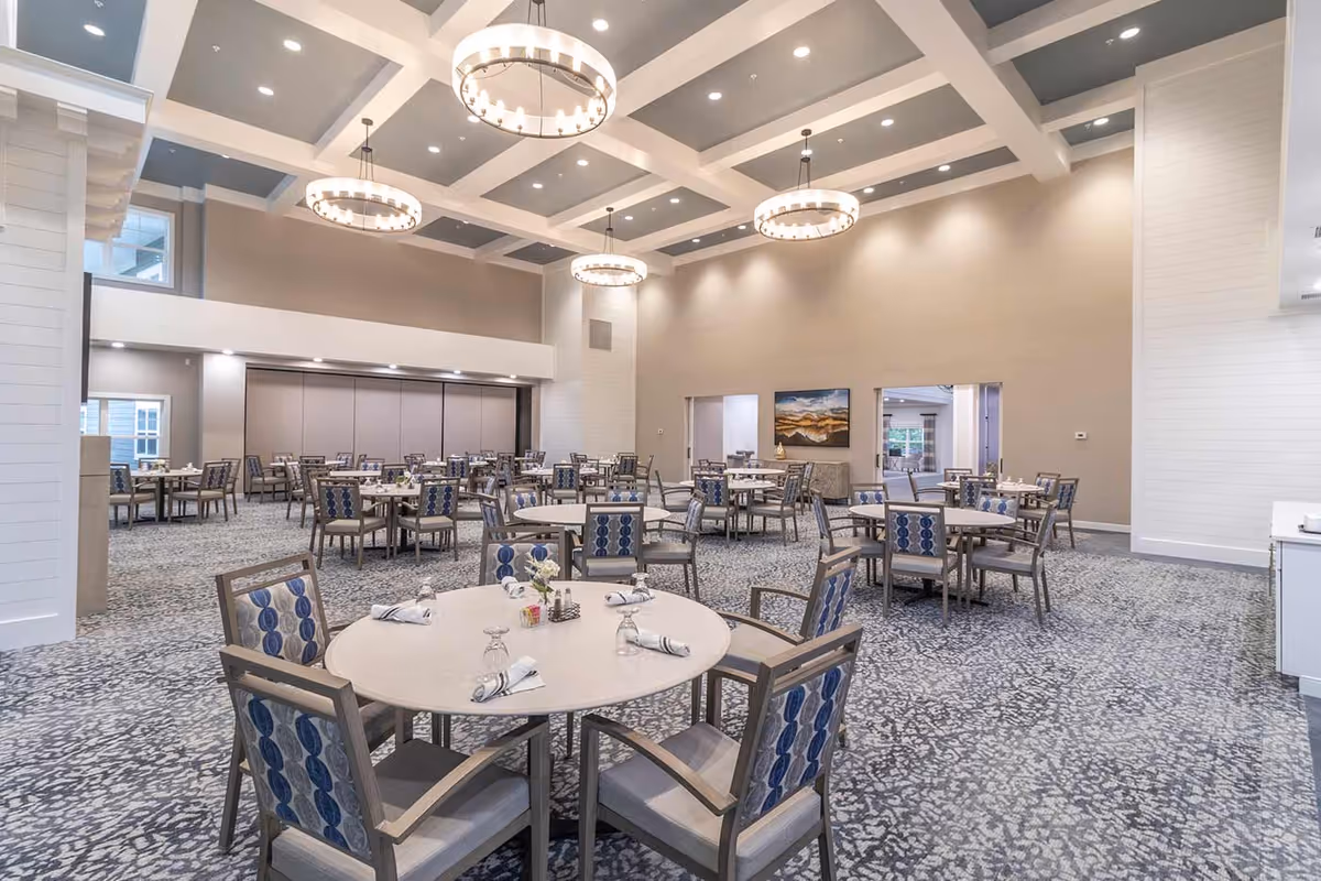 A spacious senior living dining room with multiple round tables set with napkins, glasses, and small flower arrangements. The room features high ceilings with large circular chandeliers, patterned carpet, and neutral-colored walls. There are several chairs with blue patterned upholstery around each table.