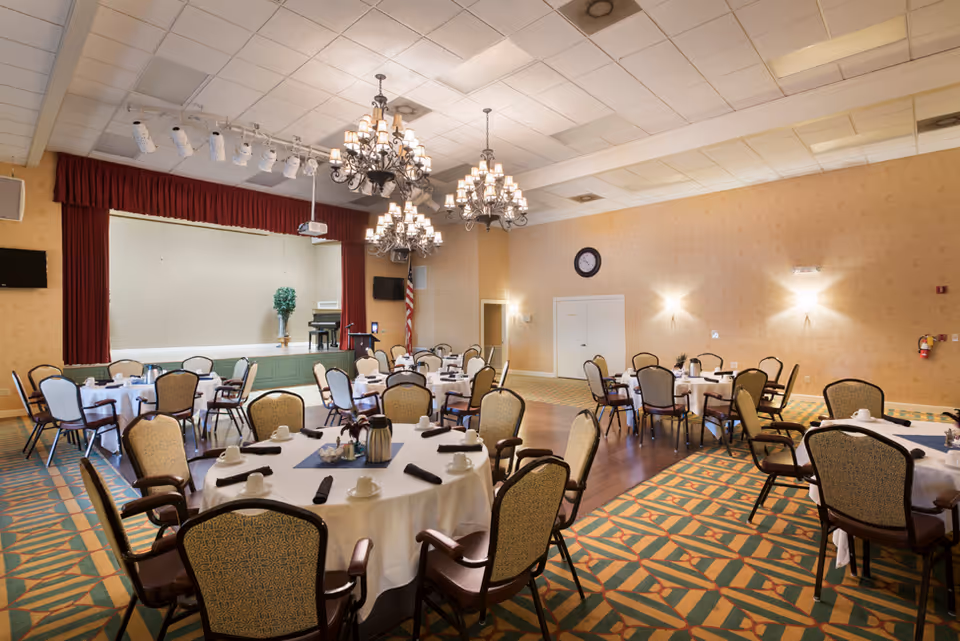 A spacious dining room with round tables covered in white tablecloths, each set with cups, saucers, and napkins. The room features patterned carpet, beige walls, and three ornate chandeliers hanging from a tiled ceiling. At the far end, there is a stage with a piano, a podium, and an American flag. Two wall-mounted televisions are visible on either side of the stage.
