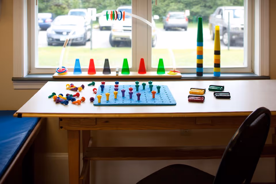 A table in a bright activity room by a window with colorful pegboard pegs, stacking cones, and small toys.