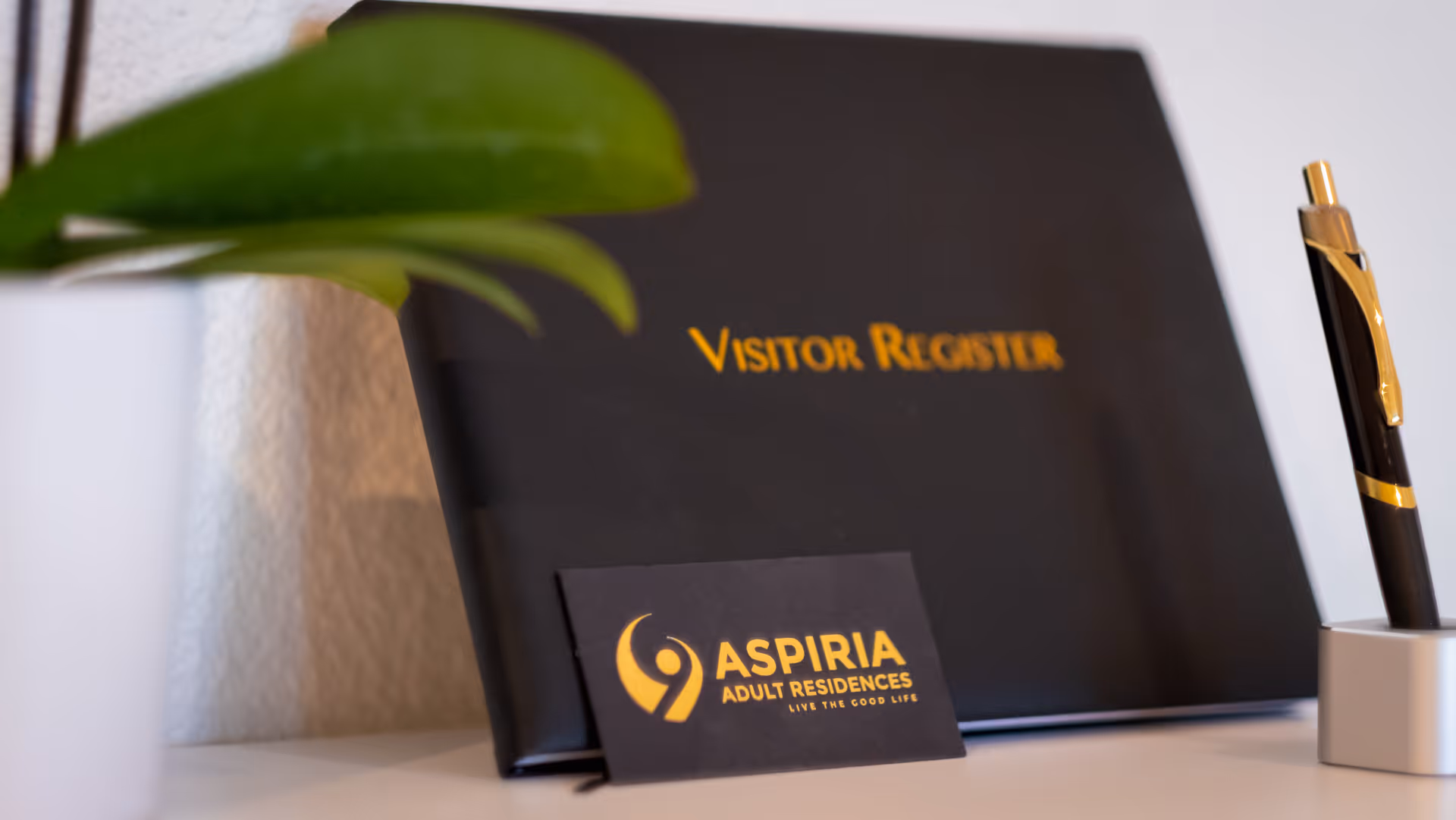 A black visitor register book with gold lettering on a white surface, accompanied by a black and gold pen in a holder and a small green plant in a white pot. A business card in front of the register displays the logo and name 'Aspiria Adult Residences' with the tagline 'Live the Good Life'.