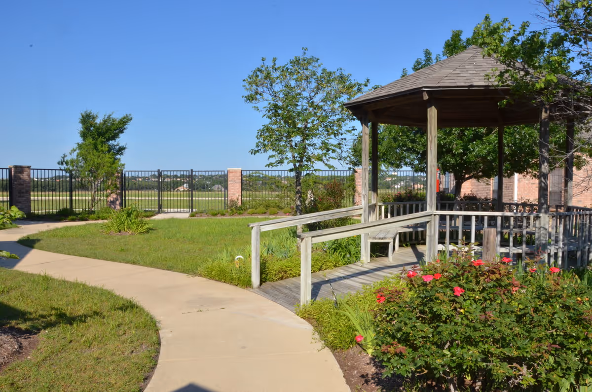Outdoor garden area with a paved walkway leading to a wooden gazebo surrounded by green grass, trees, and flowering bushes under a clear blue sky.