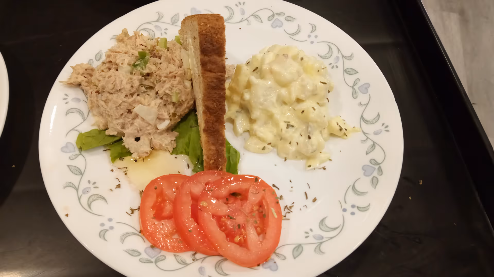 A white plate with a floral pattern containing a serving of tuna salad on a bed of lettuce, a slice of toasted bread, three slices of fresh tomato, and a portion of potato salad with herbs.