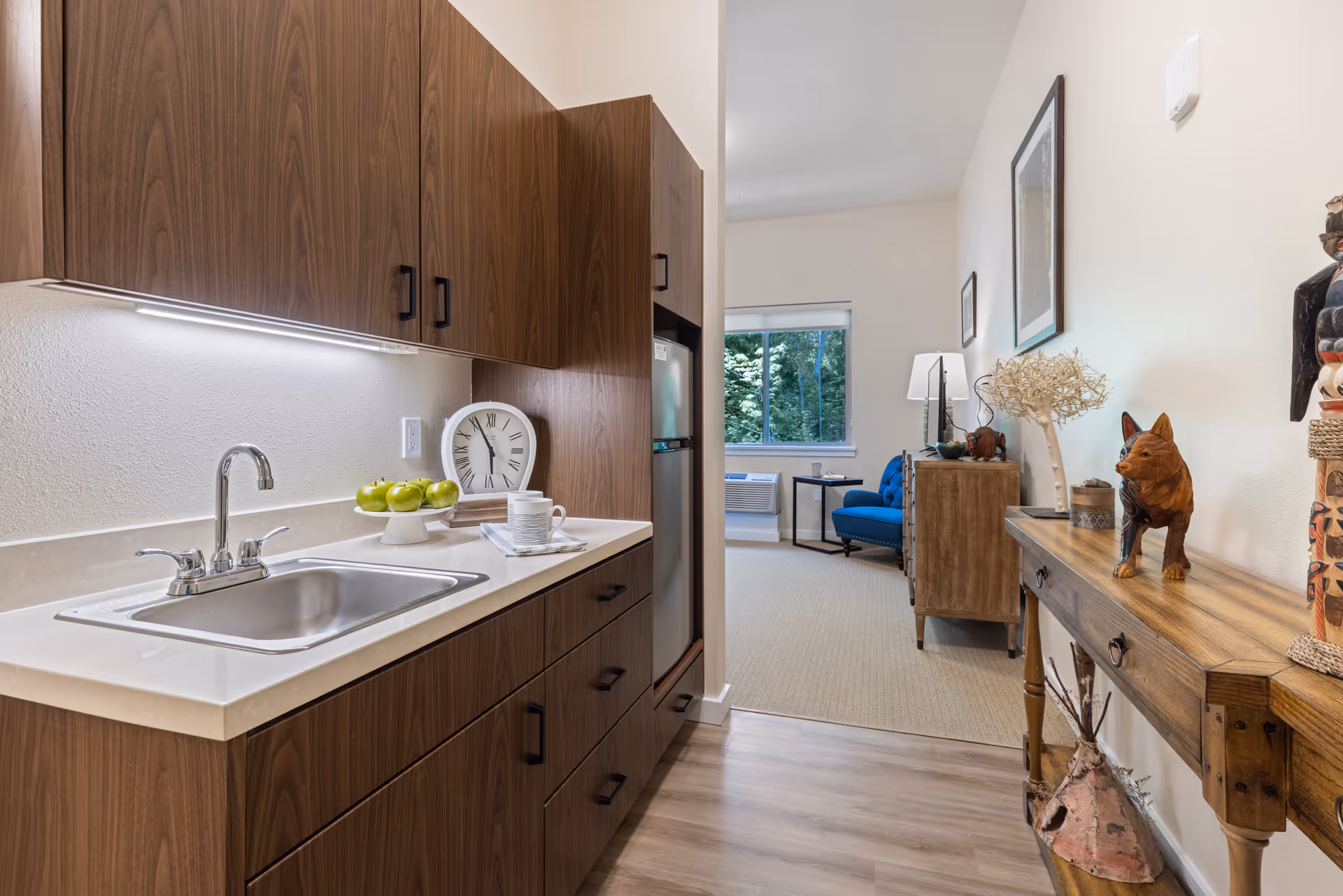A modern kitchenette with wooden cabinets, a stainless steel sink, and a countertop holding a plate of green apples, a clock, and a cup. The kitchenette opens into a living area with a blue armchair, a wooden dresser, a window with a view of greenery outside, and a wooden console table with decorative items including a small dog sculpture and a lamp.