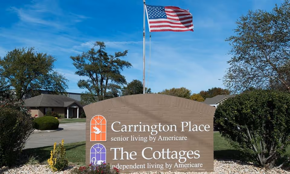 Outdoor view of a sign for Carrington Place Senior Living and The Cottages Independent Living by Americare, with an American flag flying on a pole behind it, surrounded by trees and bushes under a blue sky.