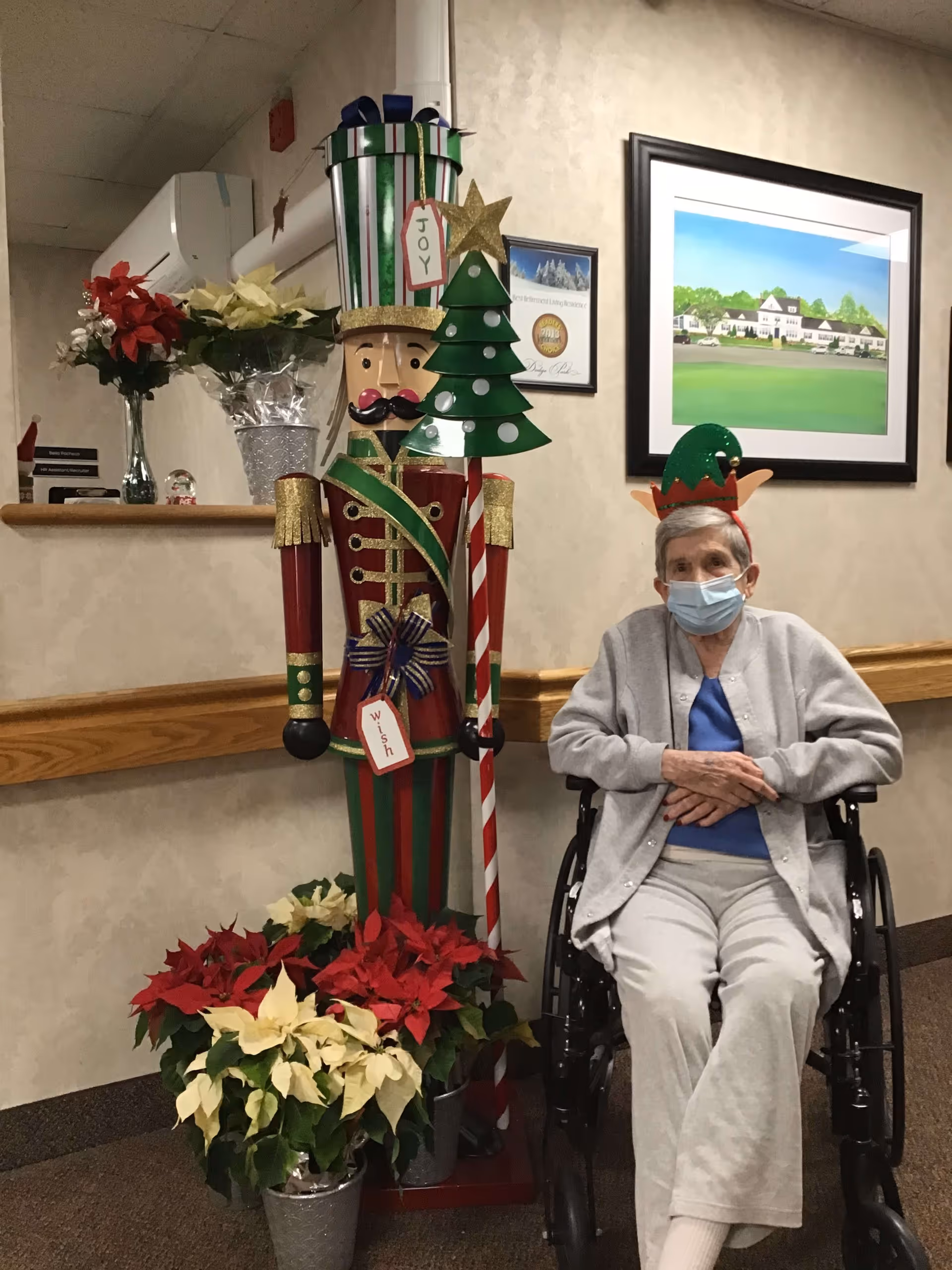 An elderly woman wearing a face mask and a festive elf hat sits in a wheelchair next to a large decorative nutcracker figure holding a Christmas tree. The nutcracker is surrounded by red and white poinsettia plants. The setting appears to be an indoor hallway with beige walls, a wooden handrail, and framed artwork on the wall.