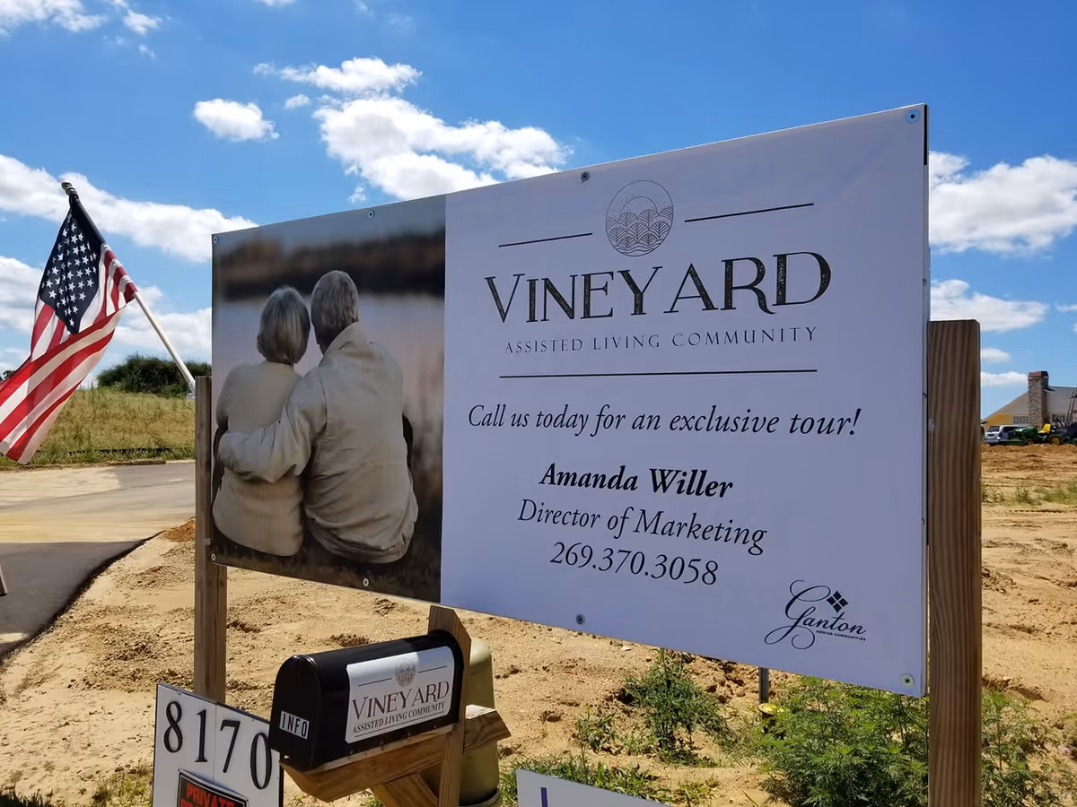 Outdoor sign for Vineyard Assisted Living Community showing a photo of an elderly couple, contact information, a mailbox and an American flag on a sunny day.