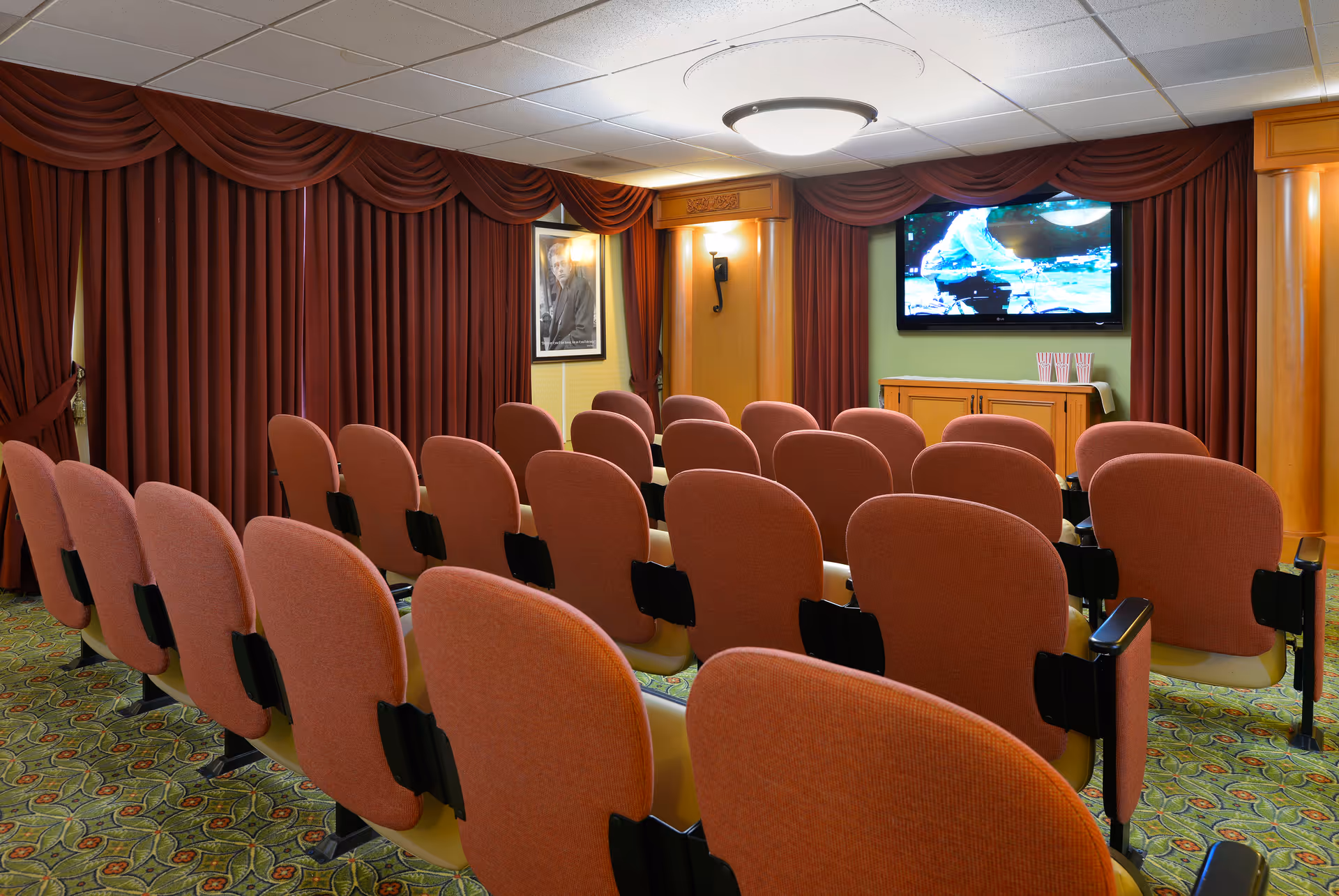 A small theater room with multiple rows of orange cushioned chairs facing a wall-mounted flat screen TV. The room has patterned carpet, maroon curtains covering the windows, and warm wood paneling with wall sconces providing soft lighting.