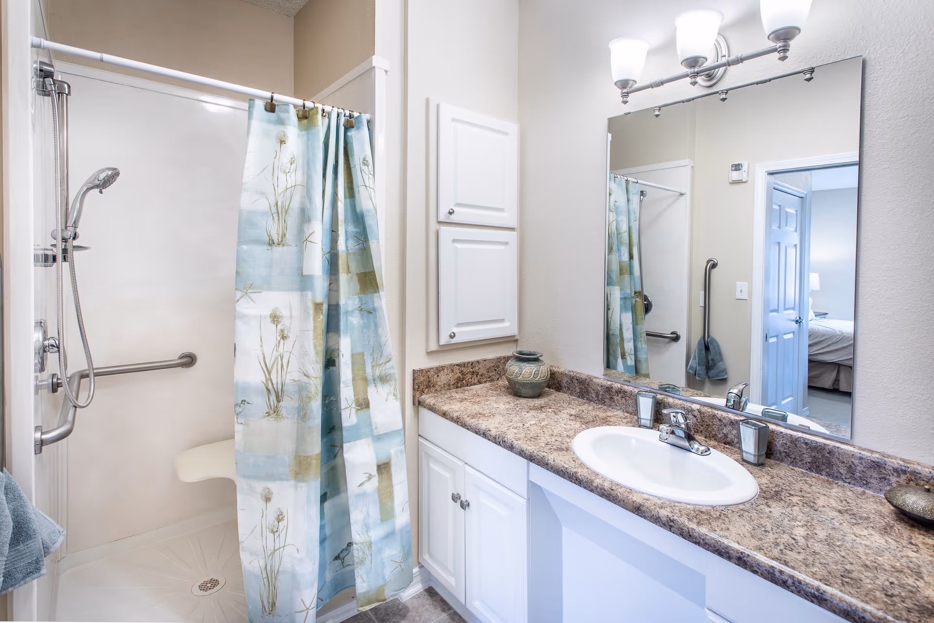A clean bathroom featuring a shower with a floral patterned curtain, a white sink with a granite countertop, white cabinets, a large mirror, and a three-light fixture above the mirror. A towel hangs on a bar near the shower, and a bedroom is visible through the open door reflected in the mirror.