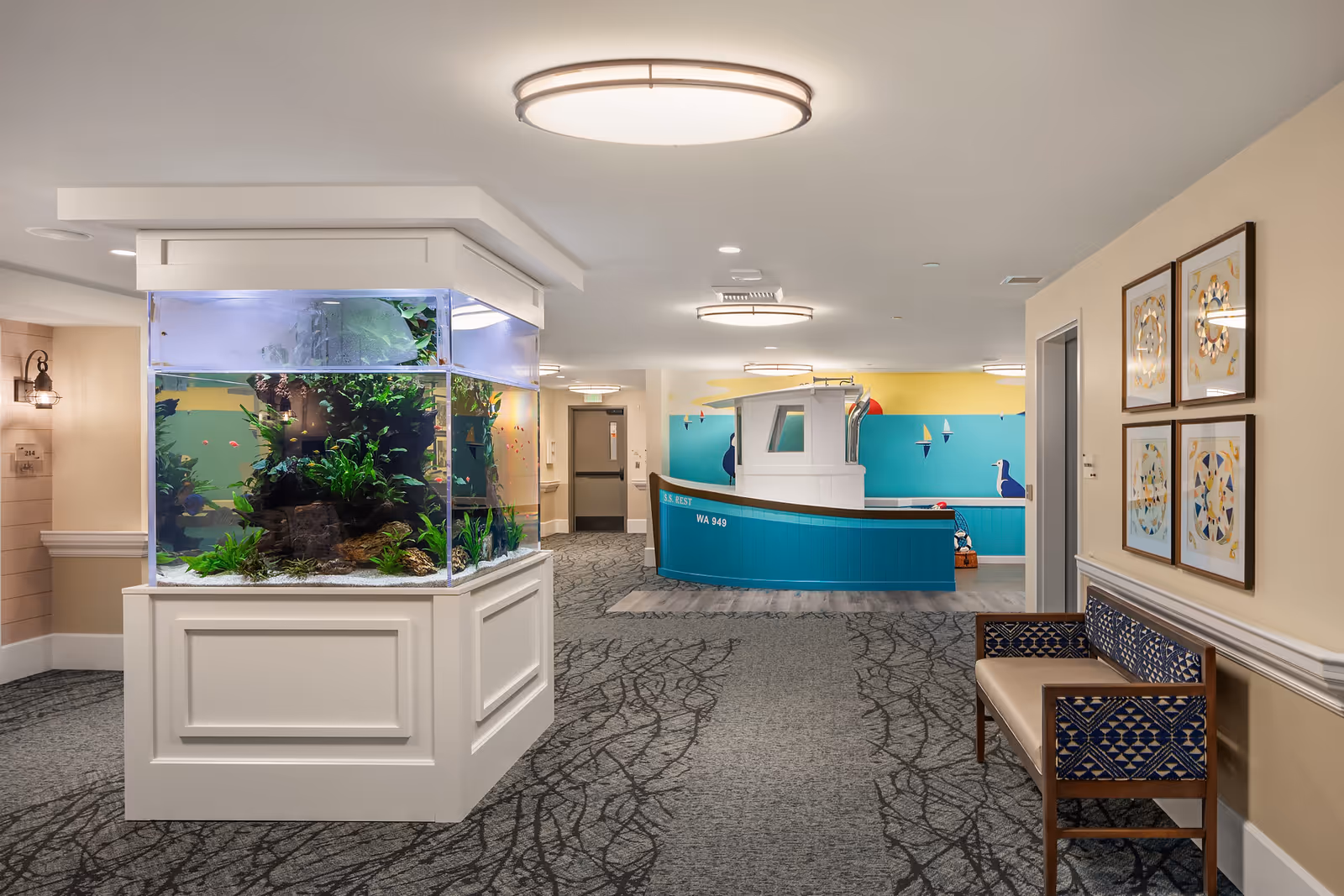 Interior hallway of Aegis Living Ballard featuring a large aquarium with plants and fish on the left, a blue and white boat-shaped play structure or seating area in the background, patterned carpet, beige walls with framed artwork, and a wooden bench with blue patterned cushions on the right.