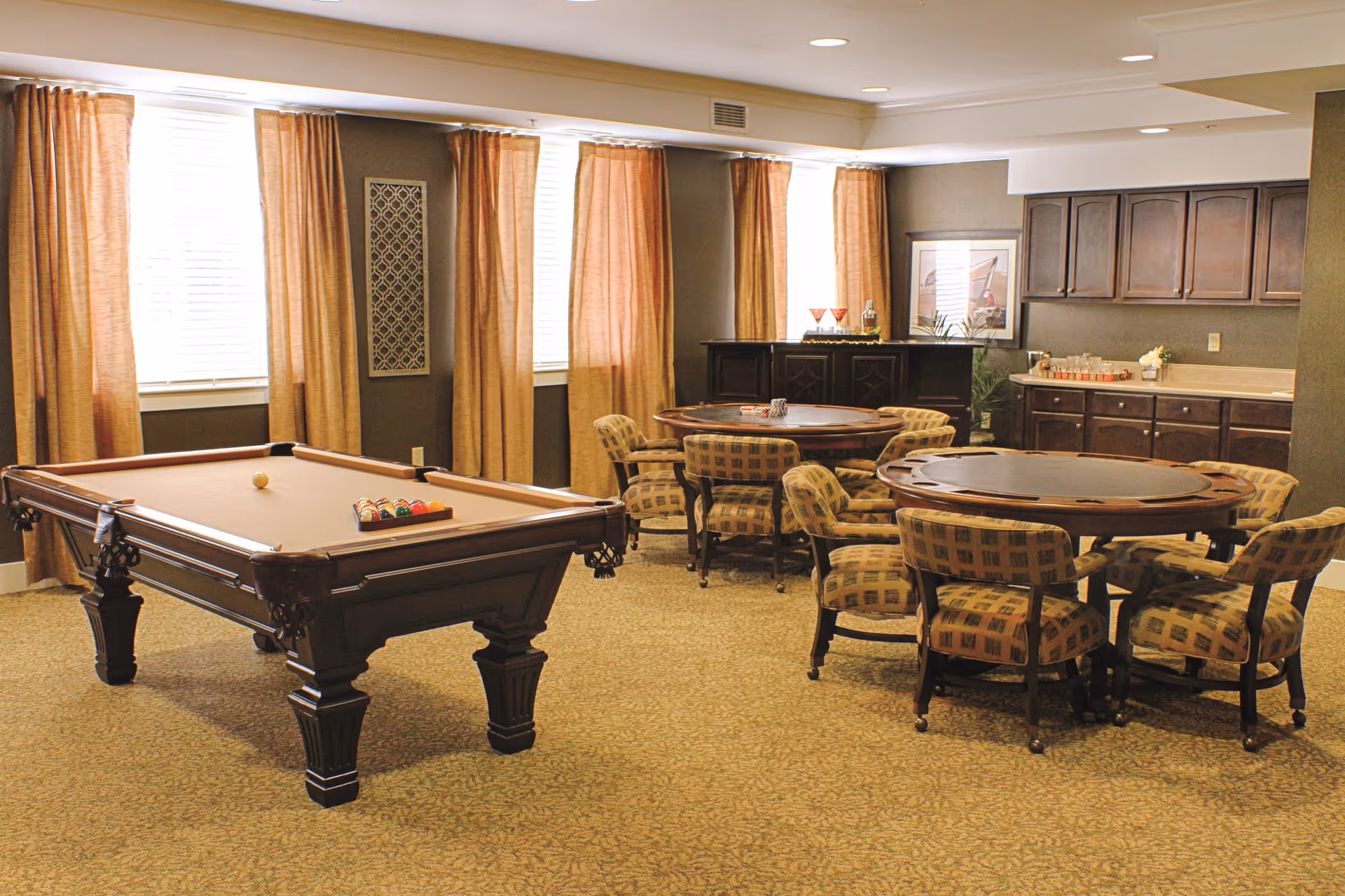 A recreational room with a pool table on the left and two round card tables surrounded by cushioned chairs on the right. The room has large windows with beige curtains, carpeted floor, and a counter with cabinets in the background.