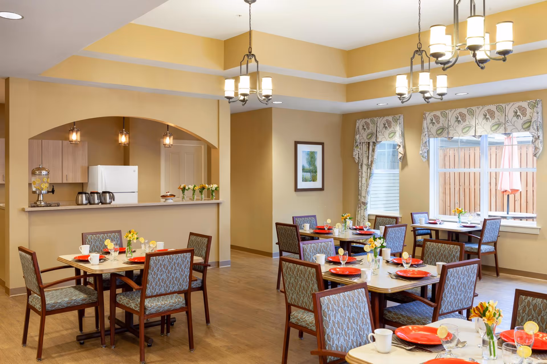A bright dining room in a senior living facility with several tables set with red plates, white cups, and glasses with lemon slices. The room features wooden chairs with patterned upholstery, large windows with floral valances, and a counter area with a refrigerator and beverage dispensers in the background. The walls are painted a warm beige color and there are hanging light fixtures providing illumination.