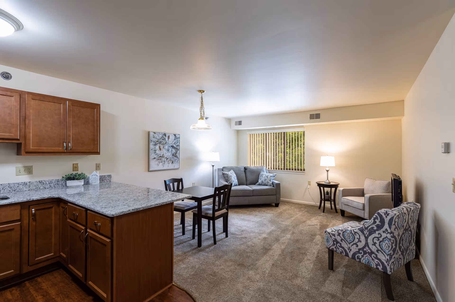 A bright and clean living area in a senior community apartment featuring a small kitchen with wooden cabinets and granite countertops on the left, a dining table with two chairs in the center, and a sitting area with a gray loveseat, two armchairs, a side table with a lamp, and a window with vertical blinds in the background.