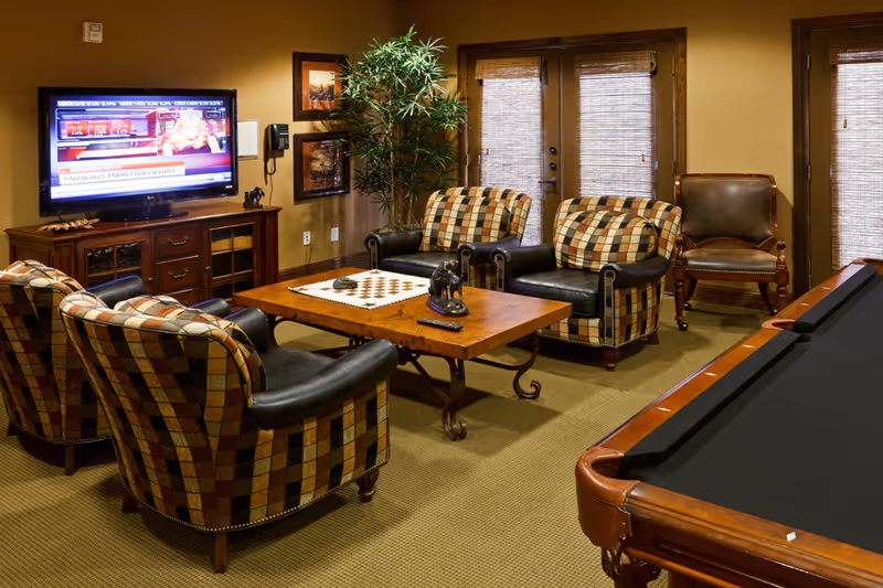 A cozy living room area with a flat-screen TV mounted on a wooden cabinet, four patterned armchairs arranged around a wooden coffee table with a chessboard on it, a potted plant in the corner, and a pool table partially visible in the foreground. There are two doors with blinds in the background.