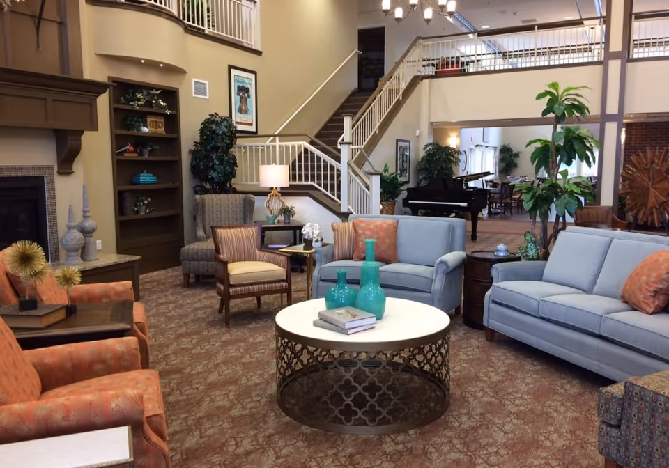 A spacious and well-decorated living room area in a senior living facility featuring comfortable seating including two blue sofas, two orange armchairs, and a wooden chair with a cushion. A round coffee table with decorative vases and books is centered in the room. There is a staircase leading to an upper level, a fireplace on the left, a bookshelf with decor items, several potted plants, and a grand piano in the background. The room has warm lighting and a patterned carpet.