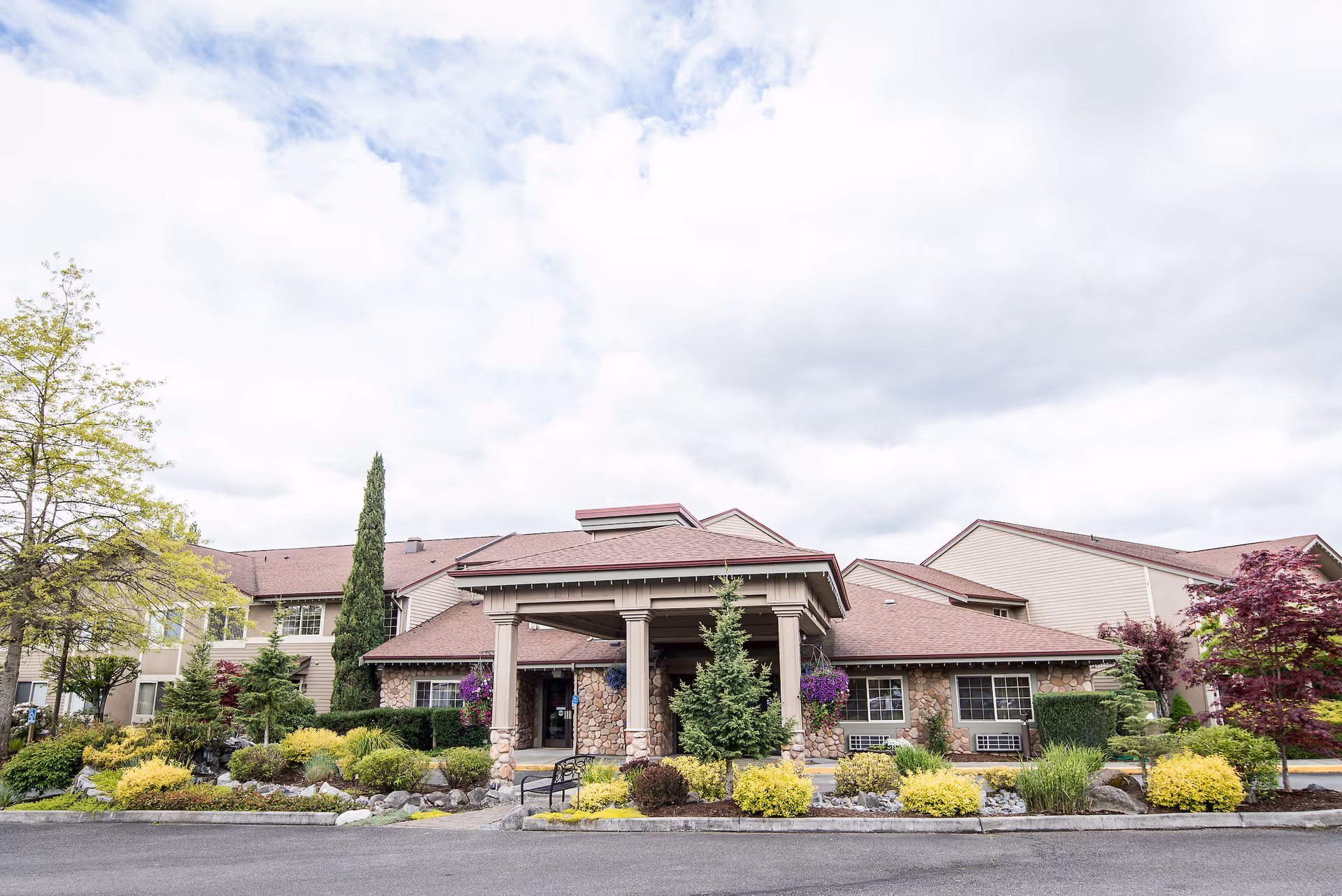 Front exterior view of a senior living facility building with a covered entrance, stone and siding facade, surrounded by landscaped bushes, trees, and flowers under a cloudy sky.