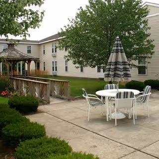 Outdoor patio area with a round white table and four white chairs with striped cushions, a large striped umbrella, a wooden gazebo, green bushes, and a two-story beige building with multiple windows in the background.