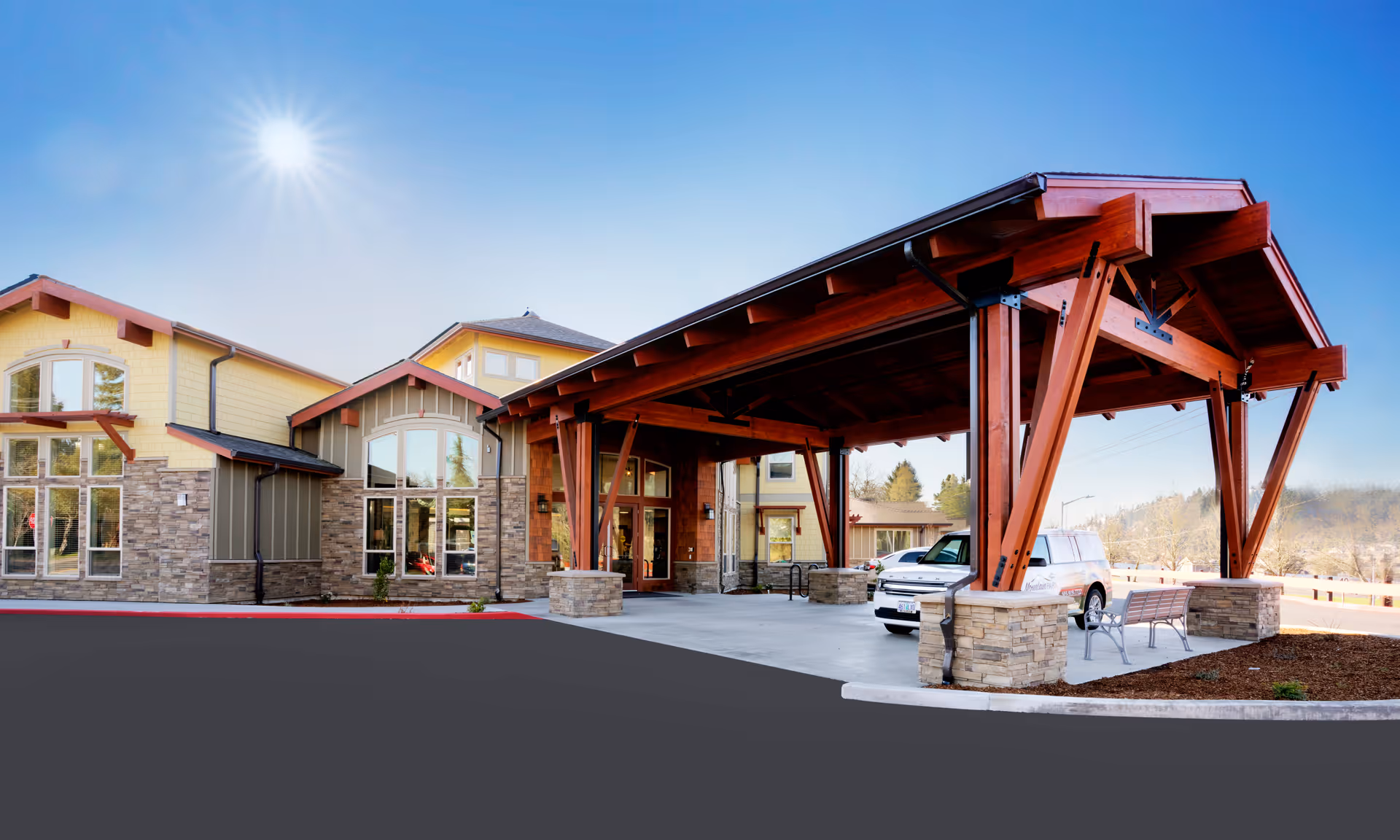 Exterior view of Mountain Park Memory Care facility showing a large covered entrance with wooden beams and stone pillars, a parked white vehicle, and a bench nearby under a clear blue sky with the sun shining.