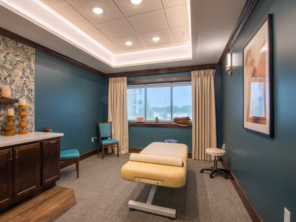 A serene therapy or massage room with a padded beige treatment table in the center, teal walls, a large window with beige curtains, two teal chairs, a small rolling stool, and a cabinet with decorative candles and a salt lamp. The ceiling has recessed lighting and a framed artwork hangs on the wall.