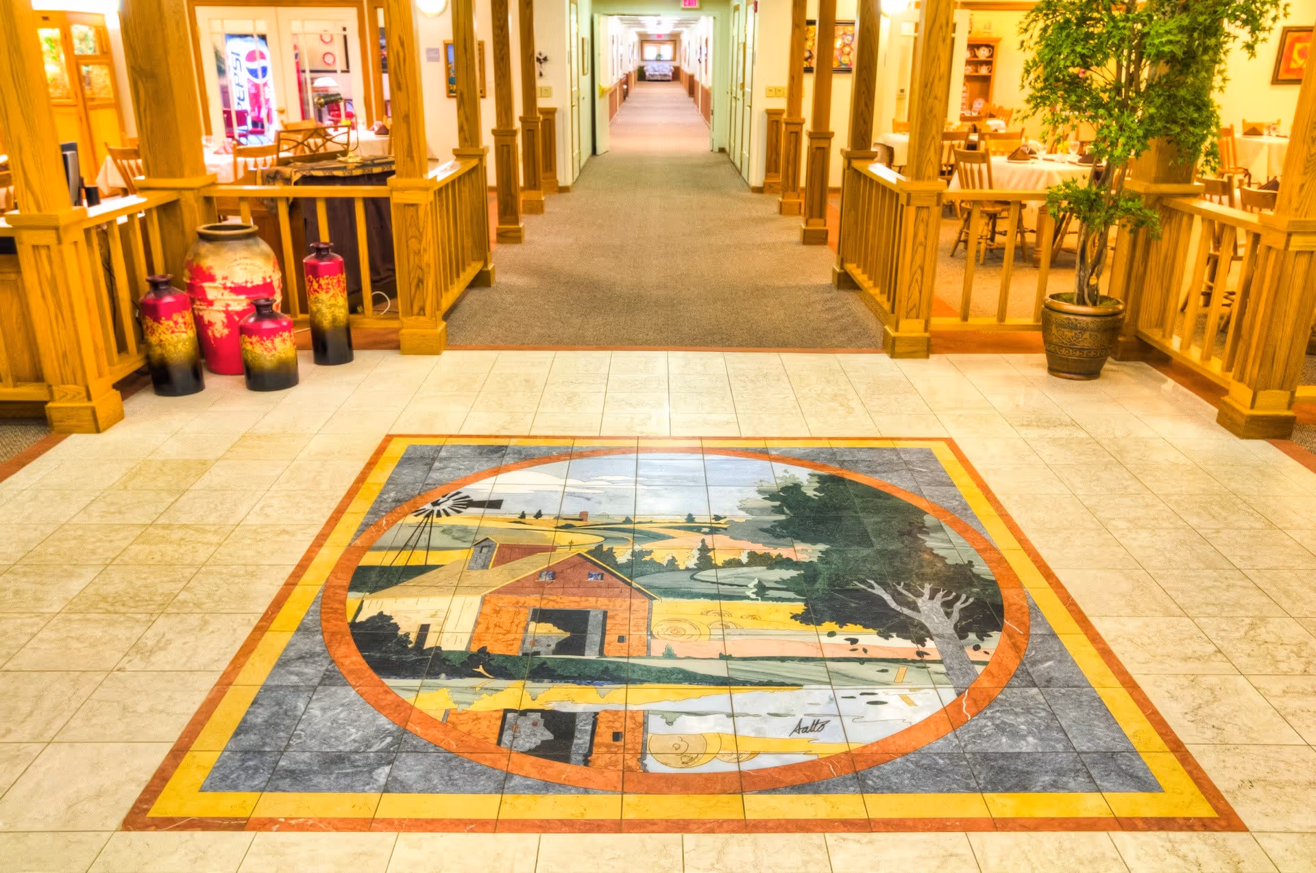 A brightly lit hallway in a senior living facility with a decorative tile floor featuring a painted rural scene of a barn, tree, and windmill. The hallway is flanked by wooden railings and columns, with seating areas and plants visible on the sides.