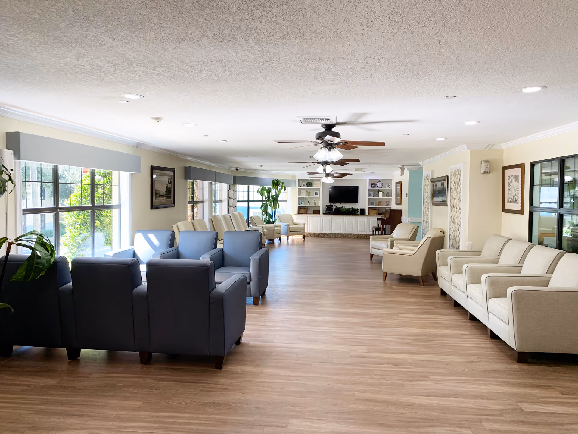 A spacious and well-lit living room area in a senior living facility with multiple armchairs arranged in small groups along the walls and near large windows. The room features wooden flooring, ceiling fans with lights, framed artwork on the walls, and a built-in entertainment center with a television and shelves at the far end.